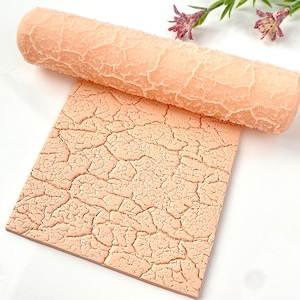 May include: A pink silicone roller with a cracked texture pattern. The roller is used to create a cracked effect on fondant or clay.