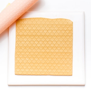 May include: A close-up of a yellow fondant sheet with a fish scale pattern, rolled out on a white surface. A pink rolling pin is lying on the surface.