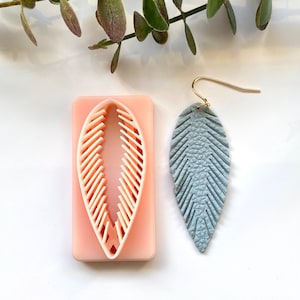 Puede incluir: Pendiente de pluma de cuero azul claro con un gancho dorado junto a un cortador de arcilla polimérica rosa en forma de pluma. El cortador tiene un diseño interior blanco.