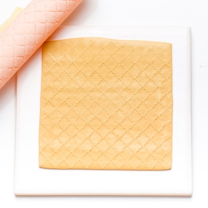 May include: A close-up of a yellow fondant sheet with a geometric pattern, rolled out on a white surface. The fondant is being used for cake decorating.