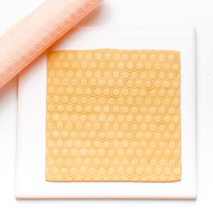 Könnte beinhalten: Nahaufnahme einer gelben Fondantplatte mit einem Wabenmuster, ausgerollt auf einer weißen Oberfläche mit einem rosa Nudelholz.