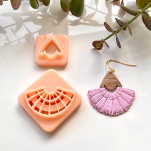 Op de afbeelding: Twee roze plastic mallen voor het maken van oorbellen, een in de vorm van een waaier en de andere in de vorm van een hart. Een paar roze kwast oorbellen met een driehoekige houten hanger.