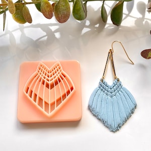 Peut inclure: Une boucle d'oreille en forme de pompon bleu clair avec un motif géométrique doré. La boucle d'oreille est présentée à côté d'un moule en silicone pour la fabrication de la boucle d'oreille.