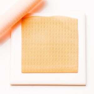 May include: A close-up of a light orange rolling pin with a chevron pattern on a white surface. The rolling pin is used to create a textured pattern on a piece of light orange fondant.