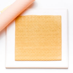 Pode incluir: Um azulejo quadrado branco com um padrão de favo de mel em relevo de cor castanho claro. Um rolo de pastelaria rosa com um padrão de favo de mel está sobre o azulejo.