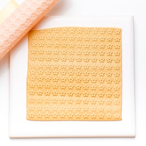 May include: A close-up of a rolling pin with a floral pattern being used to roll out a sheet of yellow fondant with a floral pattern.