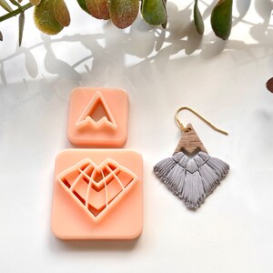 May include: Two pink plastic cookie cutters, one shaped like a mountain and the other shaped like a heart with a geometric design. A gray tassel earring with a wooden triangle pendant hangs below.