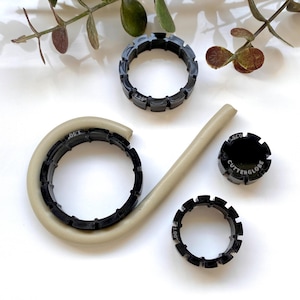May include: Four black plastic rings with different sizes and markings. The rings are on a white surface with a beige tube.