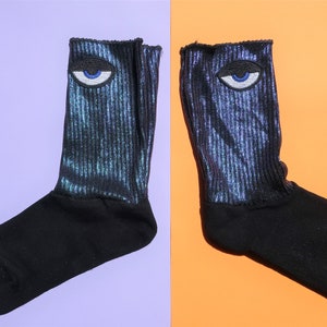 May include: A pair of black socks with a blue eye graphic on each sock. The socks are made of a sparkly material.