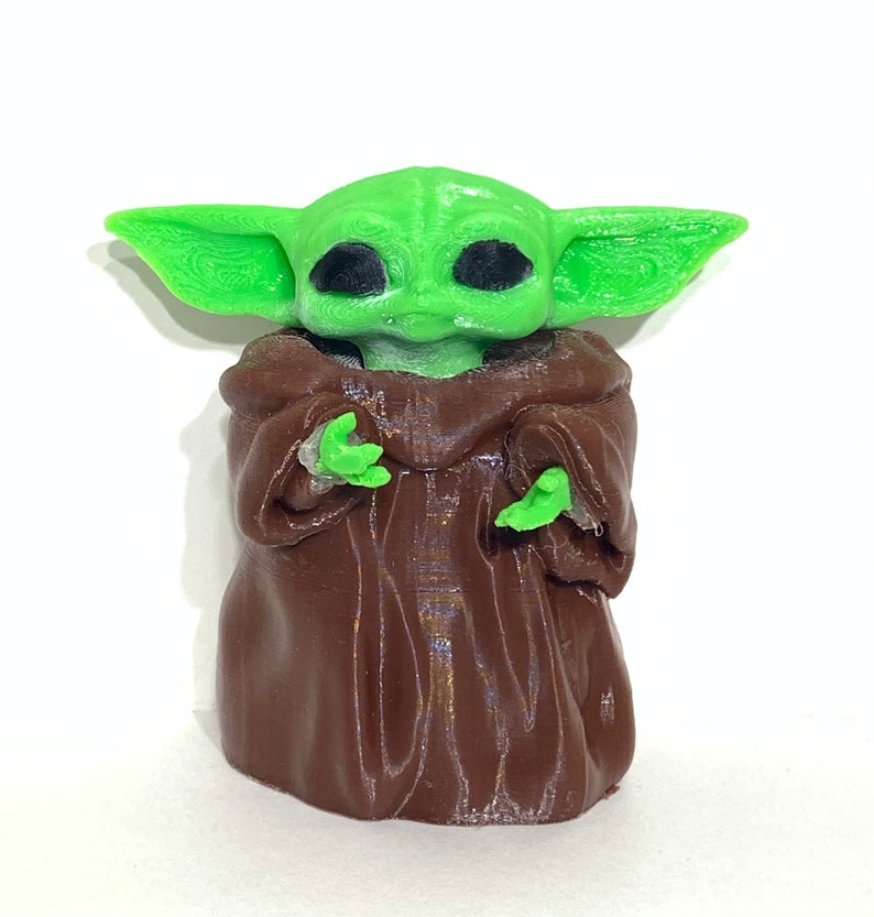 Peut inclure: Une figurine imprim&eacute;e en 3D d'un personnage de la franchise Star Wars. La figurine est de couleur verte et marron et repr&eacute;sente un personnage portant une robe marron avec de grands yeux noirs.