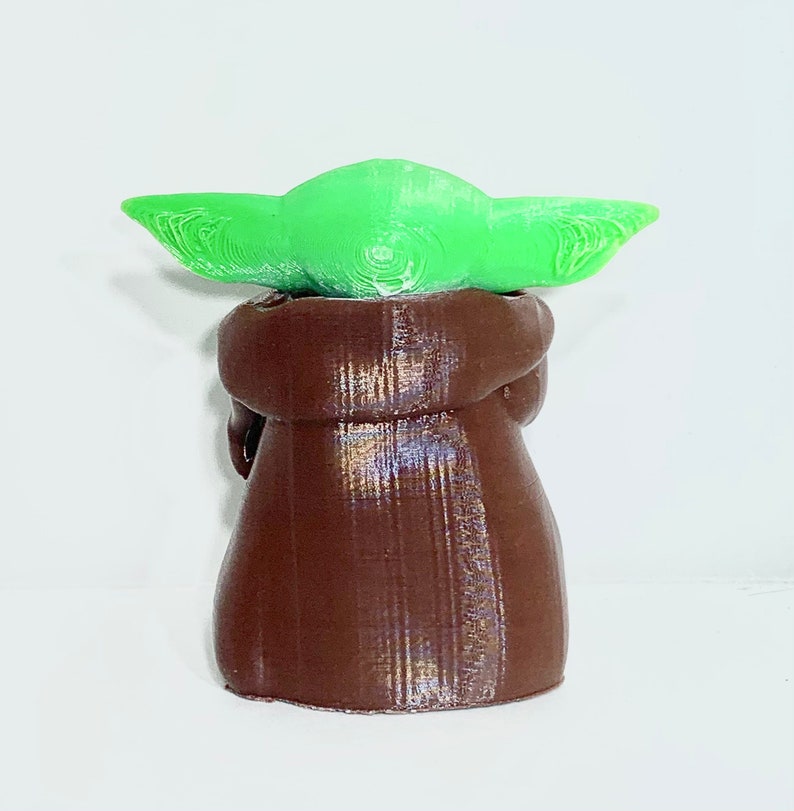 Peut inclure: Une petite figurine verte et brune d'un personnage de la franchise Star Wars. Le personnage porte une robe brune et a de grandes oreilles vertes.