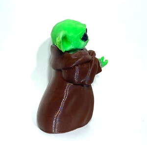 Peut inclure: Une figurine imprim&eacute;e en 3D d'un personnage vert portant une robe brune. Le personnage a de grandes oreilles et tient ses mains devant lui.