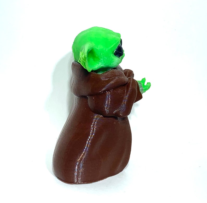 Peut inclure: Une figurine imprim&eacute;e en 3D d'un personnage vert portant une robe brune. Le personnage a de grandes oreilles et tient ses mains devant lui.