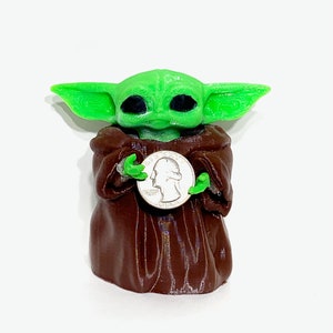 Peut inclure: Une figurine verte et brune d'un personnage de la franchise Star Wars, tenant une pi&egrave;ce de monnaie am&eacute;ricaine en argent. Le personnage a de grandes oreilles et de grands yeux.