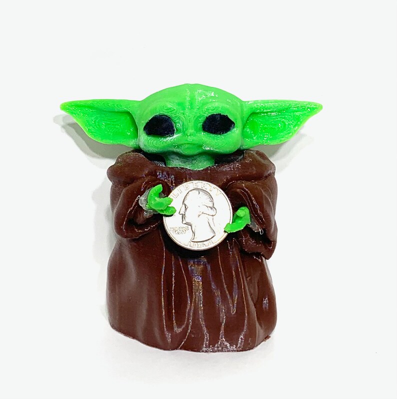 Peut inclure: Une figurine verte et brune d'un personnage de la franchise Star Wars, tenant une pi&egrave;ce de monnaie am&eacute;ricaine en argent. Le personnage a de grandes oreilles et de grands yeux.