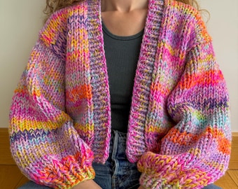 Könnte beinhalten: Ein farbenfroher, grob gestrickter Cardigan mit lockerer Passform und langen Ärmeln. Der Cardigan ist aus einer Vielzahl von leuchtenden Farben gefertigt, darunter Pink, Orange, Gelb und Blau.