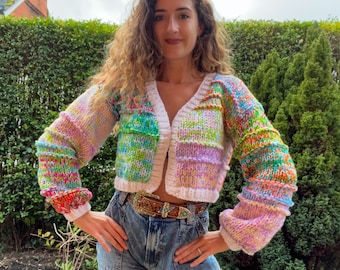 Könnte beinhalten: Ein bunter, mehrfarbiger, gehäkelter Cardigan mit weißem Besatz und V-Ausschnitt. Der Cardigan ist kurz und hat lange Ärmel. Die Person, die den Cardigan trägt, trägt auch eine blaue Jeans und einen braunen Gürtel.
