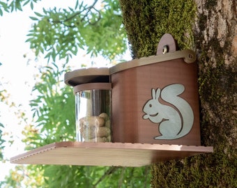 May include: A wooden squirrel feeder with a clear plastic container for nuts. The feeder is attached to a tree trunk and has a squirrel silhouette on the front.