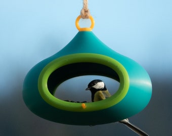 May include: A teal blue bird feeder shaped like a teardrop with a green rim and a small yellow ring at the top. A small black and white bird is perched inside the feeder.