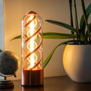 Peut inclure: Une lampe de table moderne avec une base en cuivre et un tube en verre transparent. À l'intérieur, une lumière blanche vive tourne en spirale autour d'un noyau central, avec des accents cuivrés. Un petit globe et une plante en pot sont également visibles.