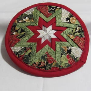 May include: A round, quilted trivet with a red border. The center features a star design in red and white, surrounded by green and black fabric with floral patterns. A decorative and functional kitchen accessory.