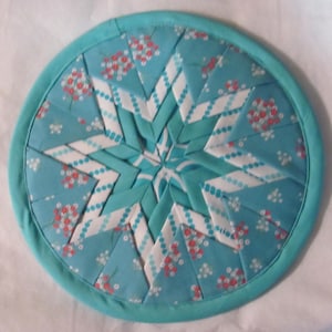 May include: A circular, quilted trivet with a turquoise border. The centre has a starburst design in white and turquoise fabric, with a floral pattern in red and white. This handmade item is ideal for protecting surfaces from hot dishes.