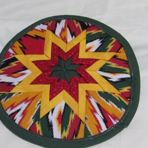 May include: A round, quilted trivet with a starburst design. The center star is made of red and yellow fabric, surrounded by a border of green, red, yellow, and white patterned fabric. The edge is finished with a dark green binding.