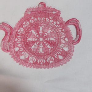 May include: A pink embroidered teapot design. The teapot features a detailed, openwork pattern, including the spout, handle, and lid. The design is set against a white background, creating a contrast that highlights the intricate details.