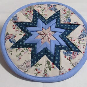 May include: A round, quilted trivet with a central star design. The star is composed of light and dark blue fabric segments, surrounded by floral patterned and polka dot fabric. The trivet has a light blue border.