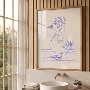 May include: A framed wall art print featuring a blue line drawing of a duck wearing a towel wrapped around its head. The duck stands on a grid pattern surface. The artwork is presented in a light-coloured wooden frame, showcasing a simple illustration.