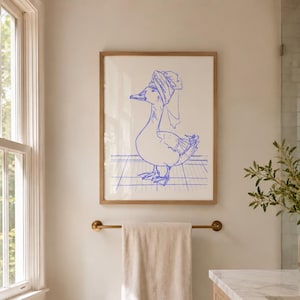 May include: A framed wall art print featuring a blue line drawing of a duck wearing a towel wrapped around its head like a turban. The duck stands on a grid-patterned floor. The artwork is enclosed in a light-coloured wooden frame.