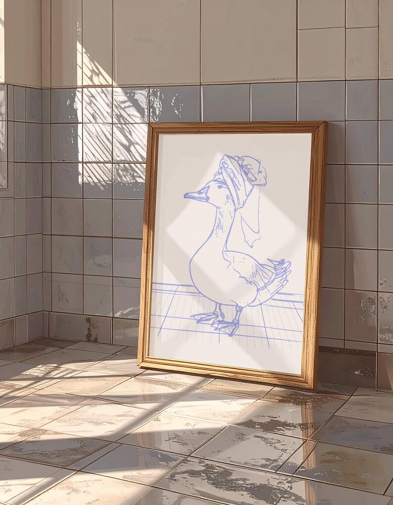 May include: A framed art print featuring a blue line drawing of a goose wearing a towel wrapped around its head like a turban. The goose stands on a grid pattern floor. The frame is made of light-coloured wood with a simple, clean profile.