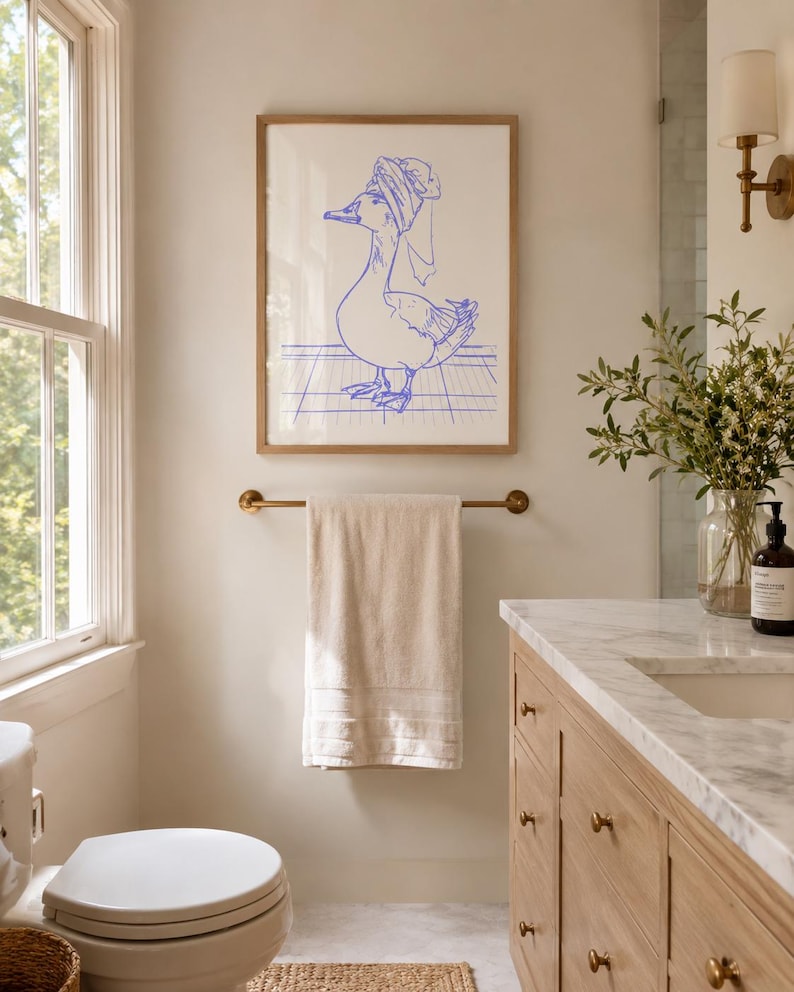 May include: A framed wall art print featuring a blue line drawing of a duck wearing a towel wrapped around its head like a turban. The duck stands on a grid-patterned floor. The artwork is enclosed in a light-coloured wooden frame.