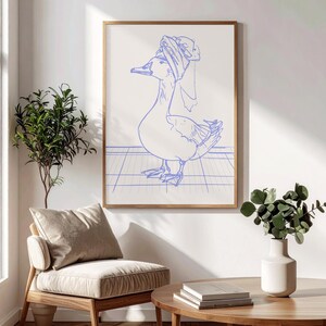 May include: A framed wall art print featuring a blue line drawing of a duck wearing a towel wrapped around its head. The duck stands on a blue grid pattern against a white background, enclosed in a thin, light-coloured wooden frame.