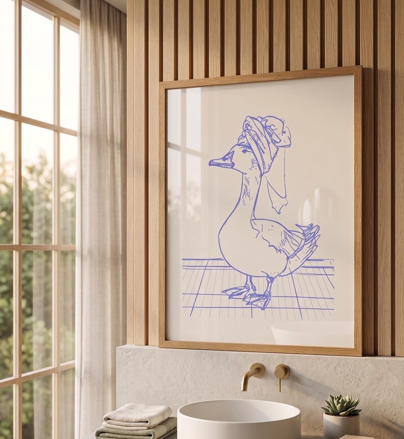 May include: A framed wall art print featuring a blue line drawing of a duck wearing a towel wrapped around its head. The duck stands on a grid pattern surface. The artwork is presented in a light-coloured wooden frame, showcasing a simple illustration.