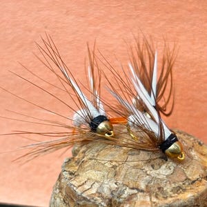 Può includere: Due mosche da pesca fatte a mano con ami e teste color oro. Le mosche hanno piume marroni e bianche e sono legate con filo nero. Sono appoggiate su un pezzo di legno, con uno sfondo color pesca.