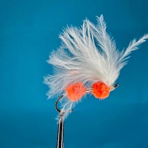 May include: Close-up of a fly fishing lure with a curved hook. The lure features two bright orange, round beads and white feathers. The background is a solid, vibrant blue.