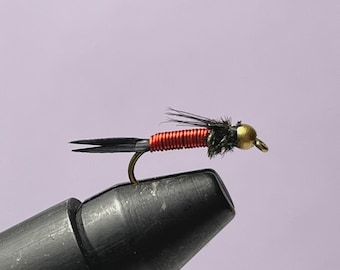 May include: A detailed image of a fishing fly, featuring a black body, red thread, and a gold-coloured head. The fly is attached to a curved hook. The background is a light purple colour. This fly is designed for angling.