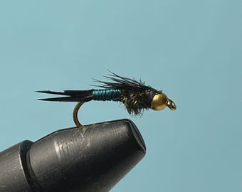 May include: A fly fishing lure with a gold head, black body, and a curved hook. The body is wrapped with teal-coloured thread. The lure is set against a light blue background. The fly is designed for fishing.