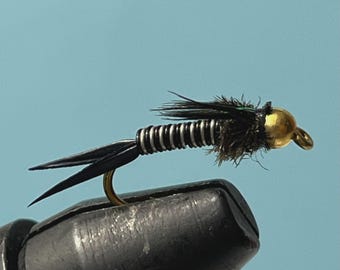 May include: A fishing fly with a gold-coloured head, a black body with silver wire wrapping, and black feather accents. The hook is gold-coloured. The fly is held in a black vice against a light blue background.