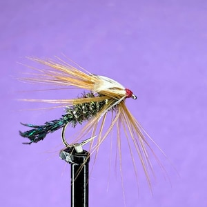 May include: A detailed fly fishing lure with a curved hook. The lure features a body of green and gold material, with tan and white feather accents. The hook is mounted on a black post against a purple background. The lure has a red head.