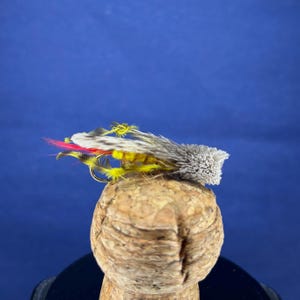 May include: A fishing fly lure featuring a yellow body, grey and white mottled wing feathers, and bright red tail fibres. The lure is constructed with a metal hook and is positioned atop a textured, light brown natural cork stopper.