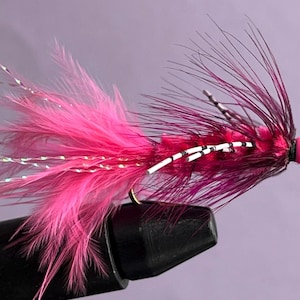 May include: A brightly coloured fishing fly in shades of pink and purple, with a gold hook. The fly has a fluffy pink body, dark purple tail feathers, and a pink bead head. White and silver accents enhance the design.