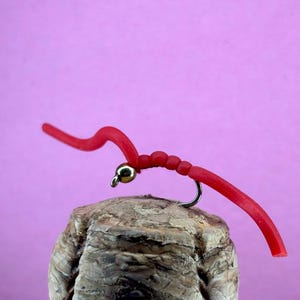May include: A red fishing fly with a curved body and a gold-coloured bead, mounted on a silver hook. The fly is positioned on a cork, set against a pink background. The fly is designed to mimic a worm.