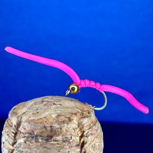 May include: A vibrant pink fishing lure with a gold bead and hook, designed to resemble a worm. It's set against a vivid blue backdrop and rests on a cork. Ideal for angling.