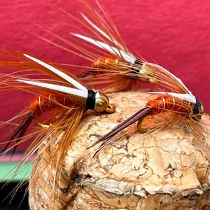 Può includere: Primo piano di tre mosche da pesca artificiali. Le mosche hanno corpi arancioni, ami dorati e ali di piume bianche e marroni. Sono appoggiate su una superficie di sughero su sfondo rosso. Attrezzatura da pesca a mosca.