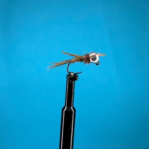 May include: A close-up of a fishing fly with a curved hook. The fly has a brown body, tan wings, and a silver bead head. The fly is attached to a black stand against a bright blue background.