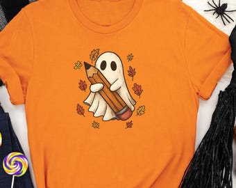 May include: Orange t-shirt featuring a cartoon ghost holding a pencil, surrounded by autumn leaves. The shirt is part of a Halloween-themed collection, with additional items like a broom, pumpkins, and candy corn.