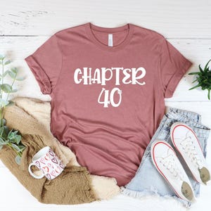 May include: A mauve t-shirt with the text "CHAPTER 40" in white lettering. The shirt is laid out on a white wooden surface with a pair of white sneakers, blue jeans, and a brown fuzzy blanket.