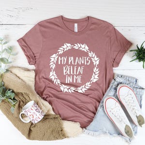 May include: A mauve t-shirt with a white graphic that says "My Plants Beleaf in Me" surrounded by a wreath of leaves.